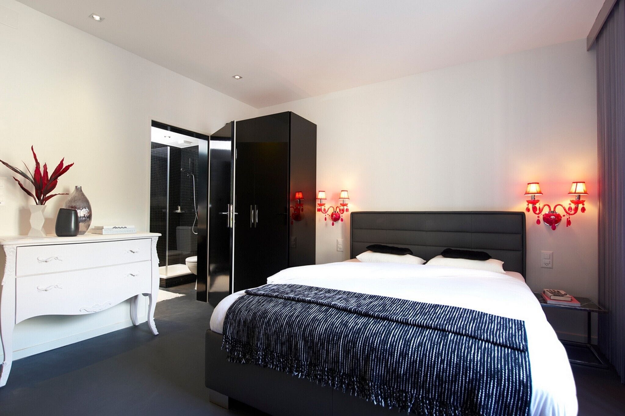 A modern hotel room with a large bed, a white dresser, a walk-in closet, and two wall-mounted lamps.