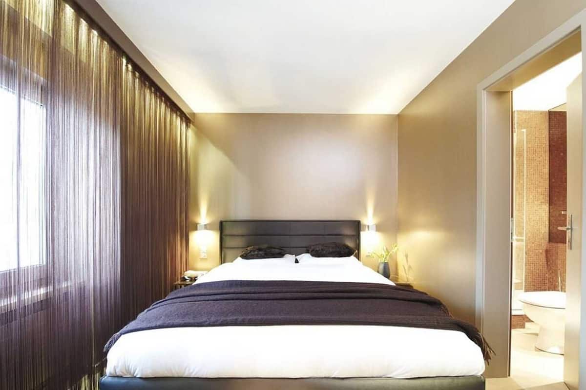 Une chambre d’hôtel avec un grand lit, deux lampes de chevet, une tête de lit sombre et une vue sur la salle de bain par une porte ouverte.