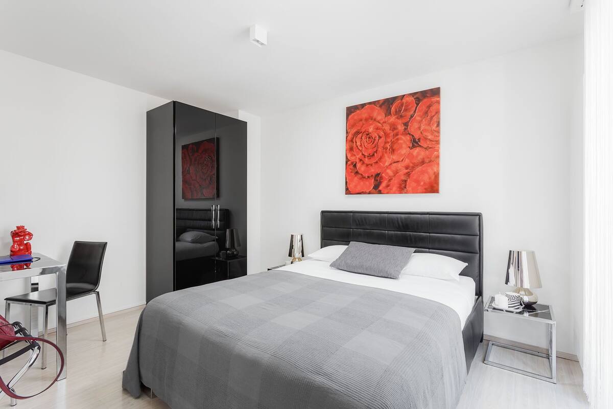 Une chambre d’hôtel moderne avec un grand lit, un bureau et une chaise. Un tableau représentant des fleurs rouges est accroché au mur.