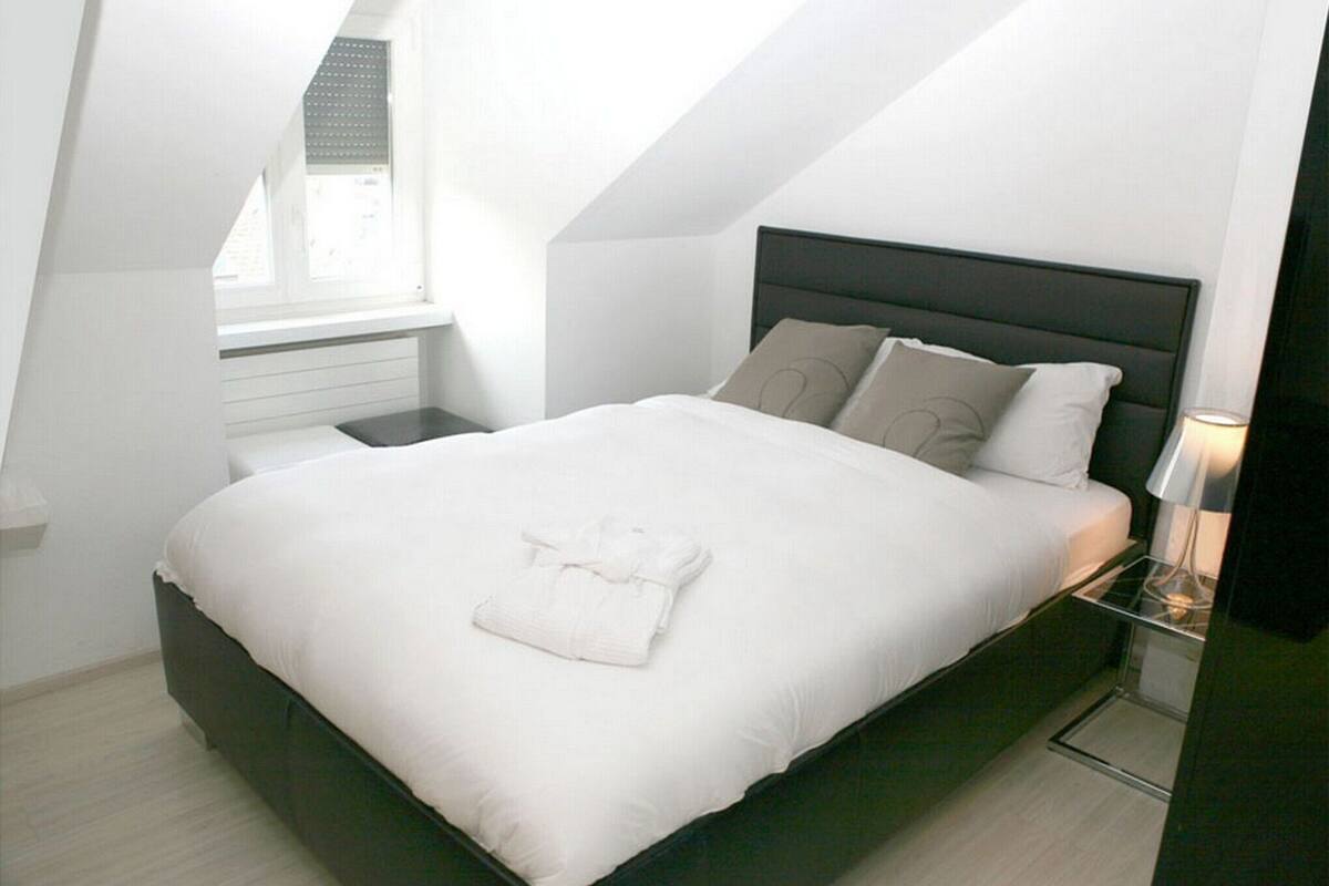 Un lit bien fait, avec du linge de lit blanc et une serviette pliée, une table de chevet avec une lampe et une fenêtre avec des stores.