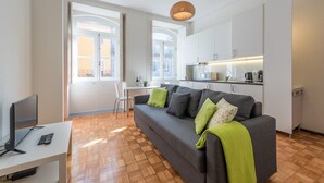 A modern living room with a grey sofa, a small white desk with a TV and remote, and a kitchen area with white cabinets and a hanging light fixture. - GuestReady - Clerigos-Virtudes Apartments (Porto)
