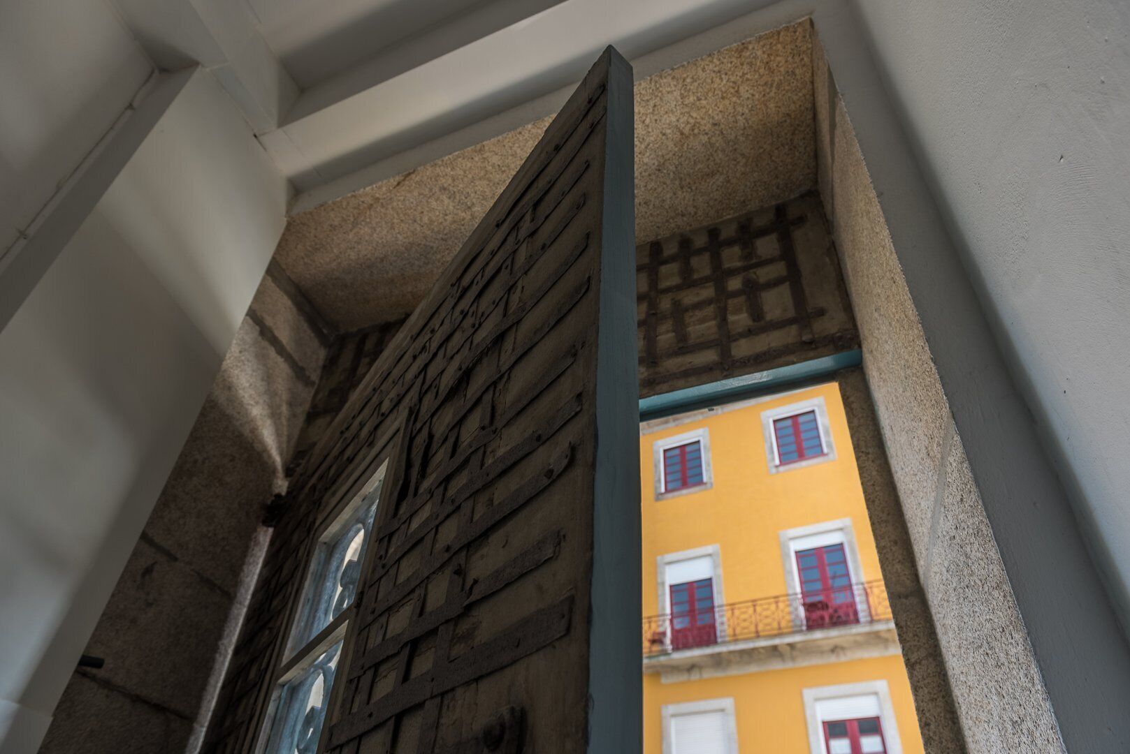 A narrow corridor with a wooden door and a view of a yellow building with red shutters.