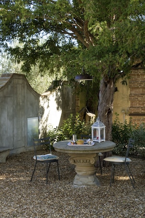 Une table ronde en pierre, accompagnée de deux chaises, d’une lanterne et d’un décor rustique agrémenté d’un grand arbre et d’un mur en briques.
