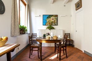 Un coin repas avec une table ronde, six chaises, une plante en pot et un tableau représentant une ville côtière accroché au mur.