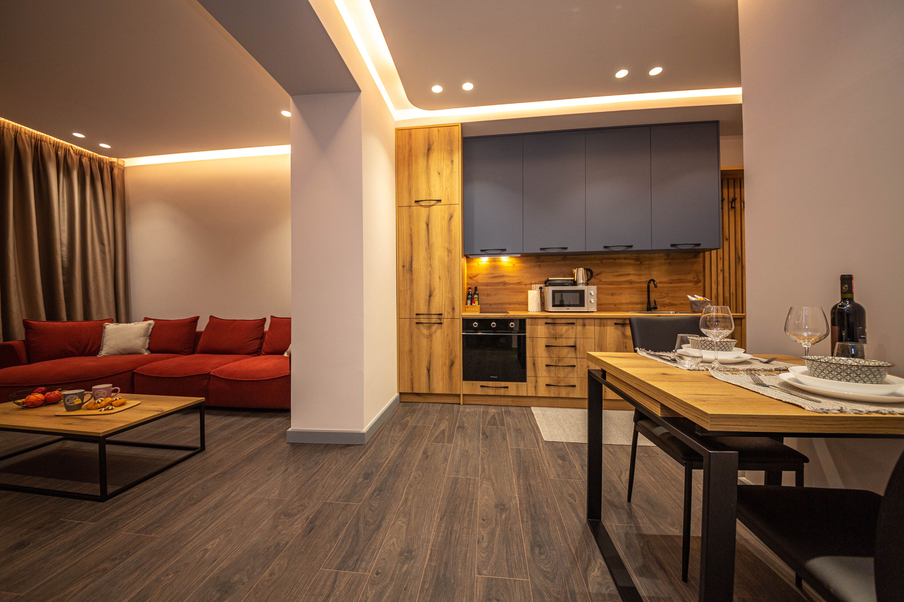 Un salon moderne avec un canapé rouge, une table à manger en bois dressée pour deux personnes et un coin cuisine doté de meubles et d’appareils électroménagers en bois.