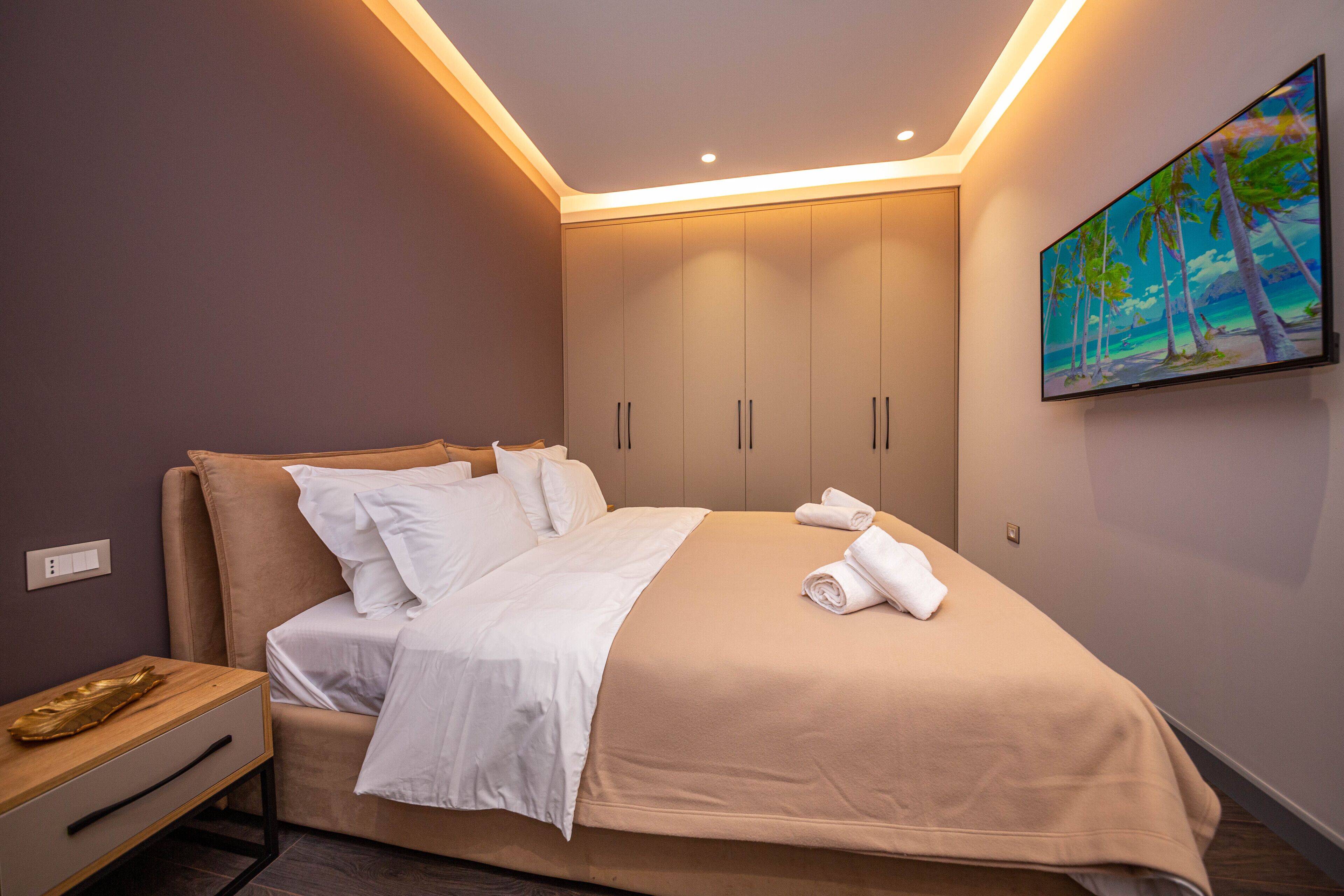 Una cama bien hecha con sábanas blancas, una mesita de noche de madera y un cuadro con una playa en la pared.