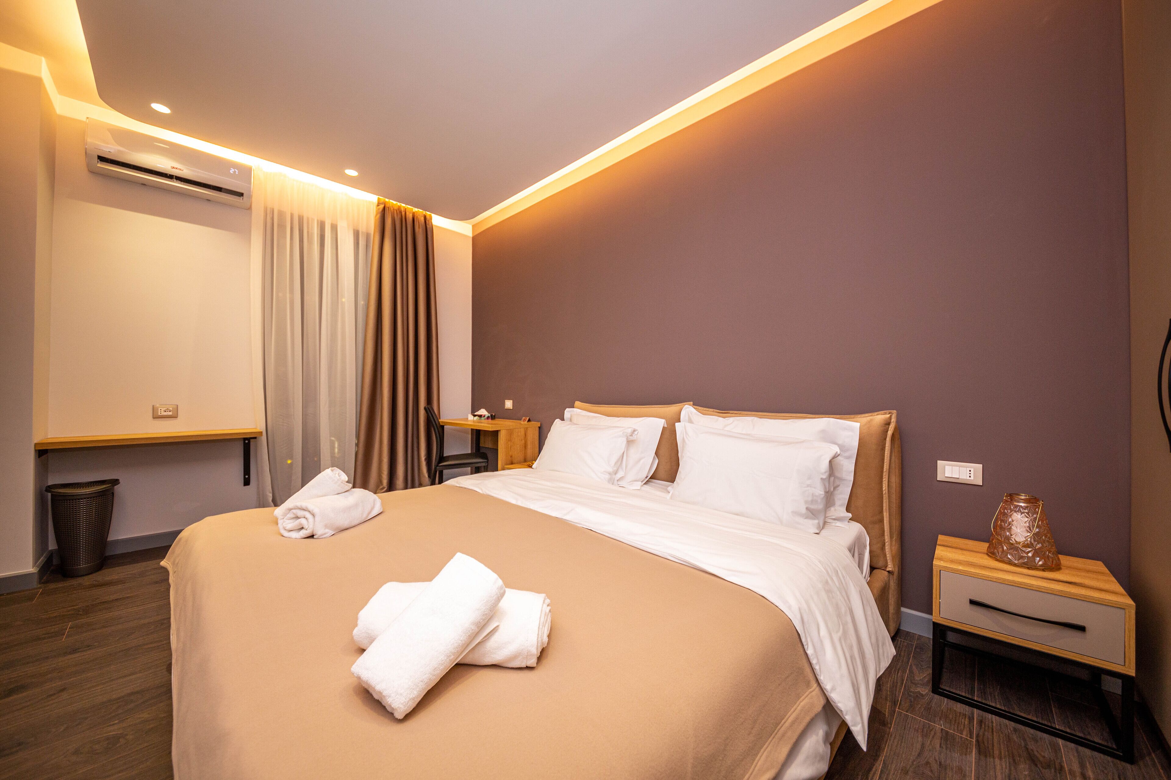 A hotel room with a large bed, a bedside table, a nightstand, and a wall-mounted air conditioner.