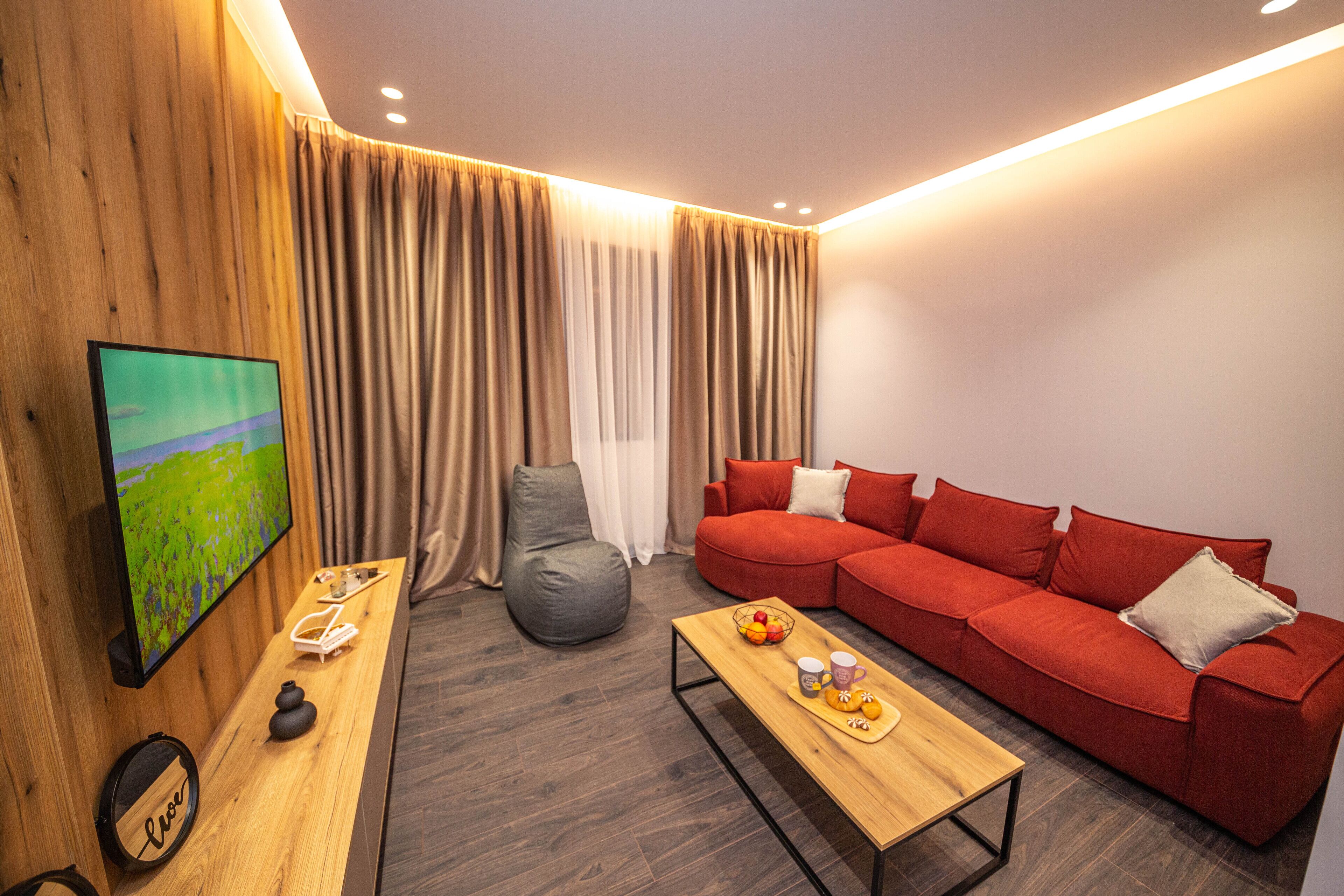 Ein modernes Wohnzimmer mit einem roten Sofa, einem Holztisch, einem grauen Sitzsack und einem Fernseher an der Wand.
