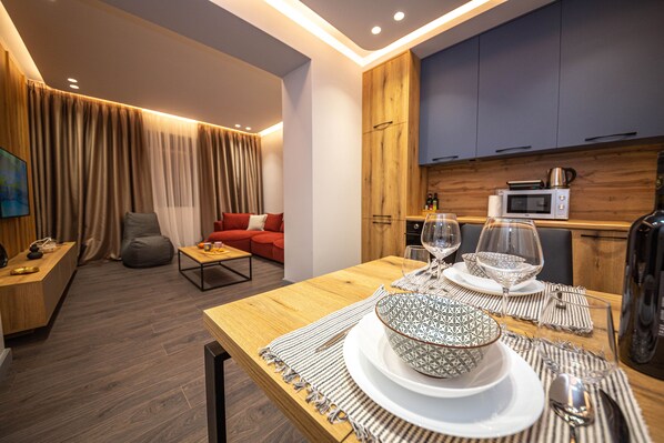 Uma sala de estar moderna com uma mesa de jantar de madeira posta para dois, um sofá vermelho e uma cozinha equipada com micro-ondas e chaleira.