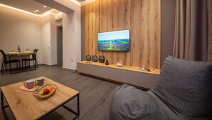 A modern living room with a flat-screen TV mounted on a wooden wall, a grey sofa, a wooden coffee table with fruit, and a dining area in the background. - Blloku Unique Apartments (Tirana)