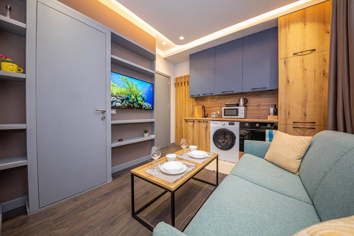 A compact living space with a sofa, a dining table set for two, a washing machine, a microwave, and a TV on a wooden cabinet wall.