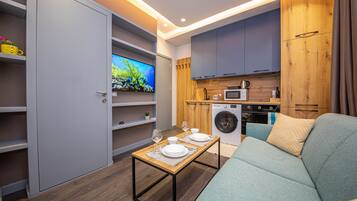 A compact living space with a sofa, a dining table set for two, a washing machine, a microwave, and a TV on a wooden cabinet wall.