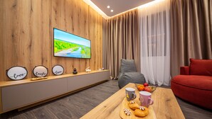 A modern living room with a wooden wall, a flat-screen TV, a grey sofa, a red sofa, a wooden coffee table with pastries and a fruit bowl, and a window with curtains. - Blloku Unique Apartments (Tirana)