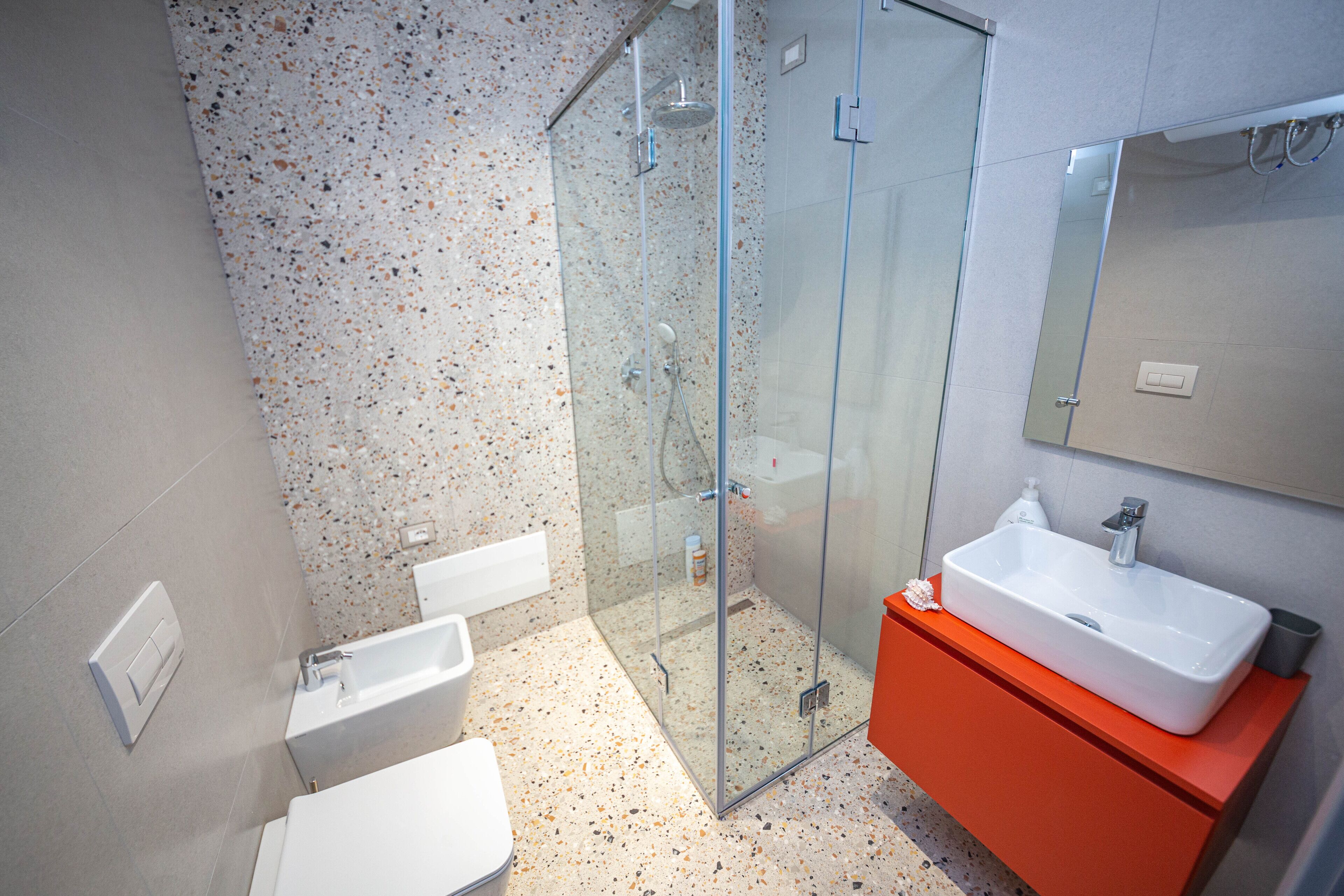 Un baño moderno con cabina de ducha de vidrio, inodoro blanco y tocador naranja con lavabo blanco.