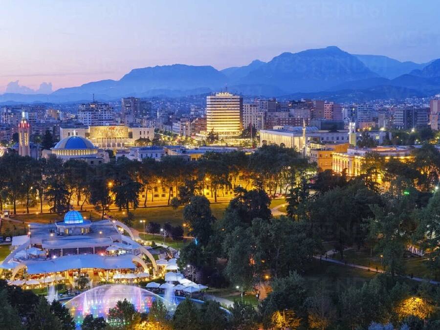 A cityscape at dusk with illuminated buildings, a prominent dome structure, and mountains in the background.