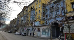 Uma fileira de edifícios altos com varandas, carros estacionados e pedestres na rua.