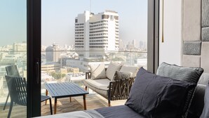 A modern living room with a sofa, armchair, and table, overlooking a cityscape with tall buildings. - MAMAD High End 2BR with City & Sea View by FeelHome (Tel Aviv - Jaffa)