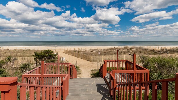 Een houten vlonder die naar een strand leidt, met een heldere lucht en wolken.