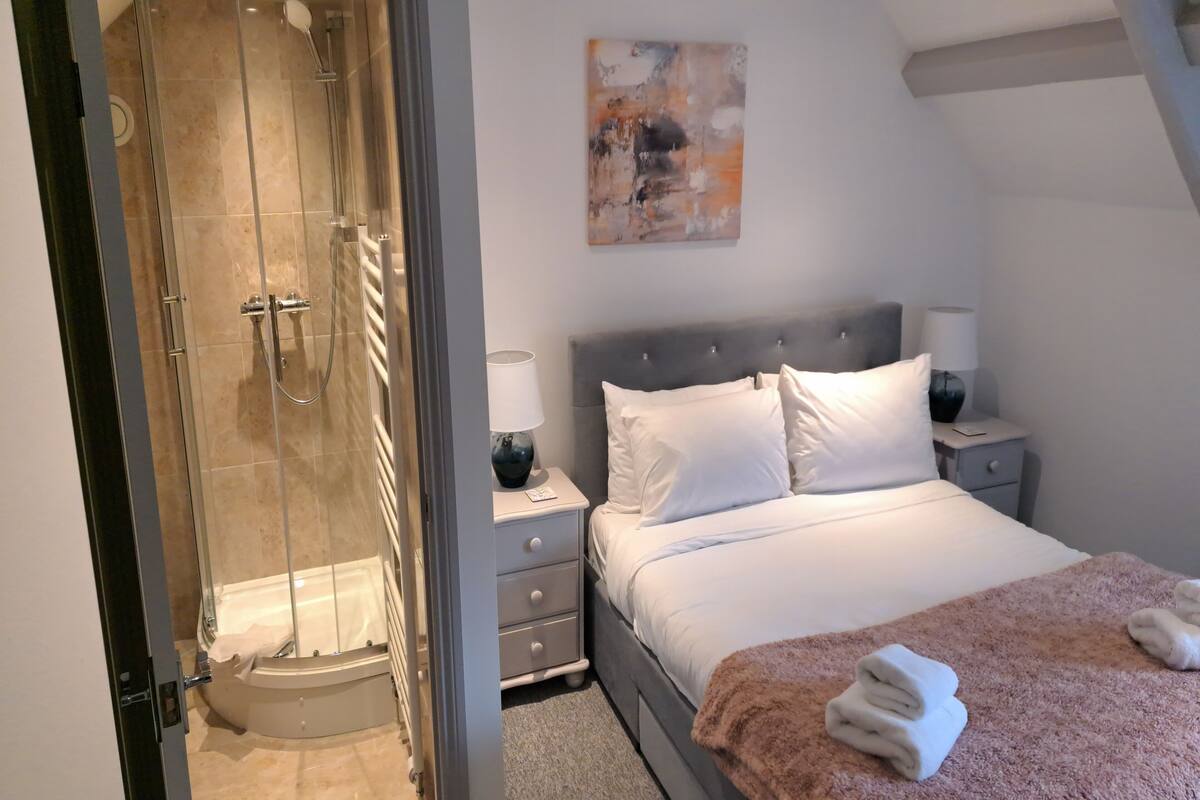 A bedroom with a glass-enclosed shower, a bed with white bedding, a nightstand with a lamp, and a painting on the wall.