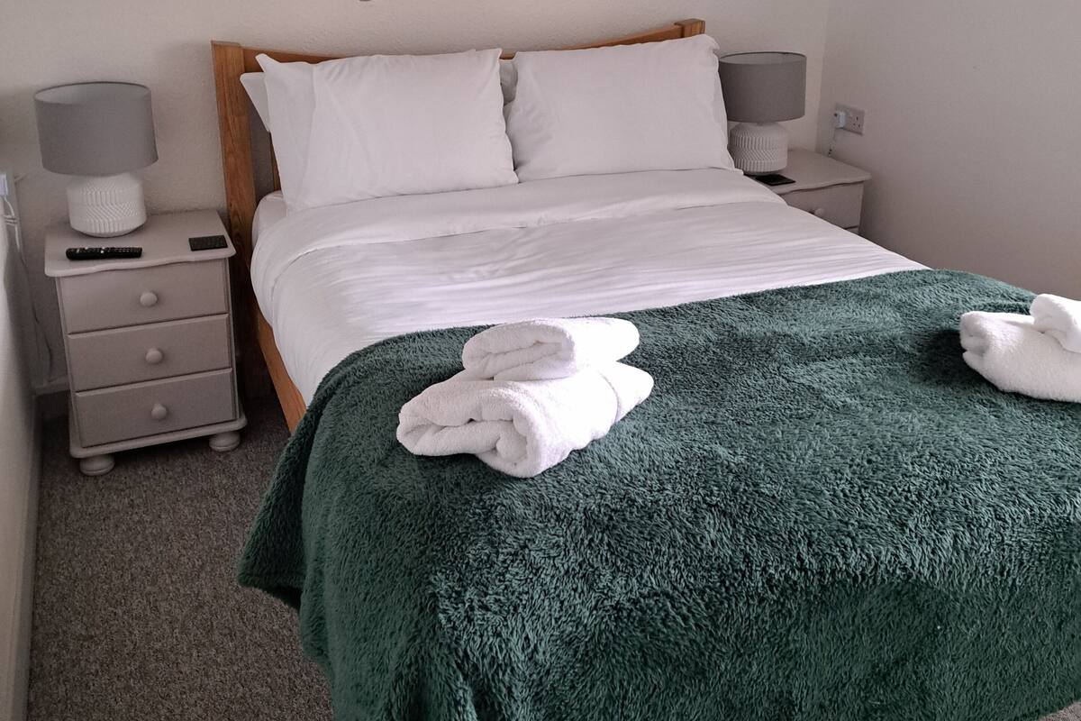 A neatly made bed with white linens, a green blanket, and two folded towels on a dark green carpet. There are two bedside tables with lamps and a small drawer.