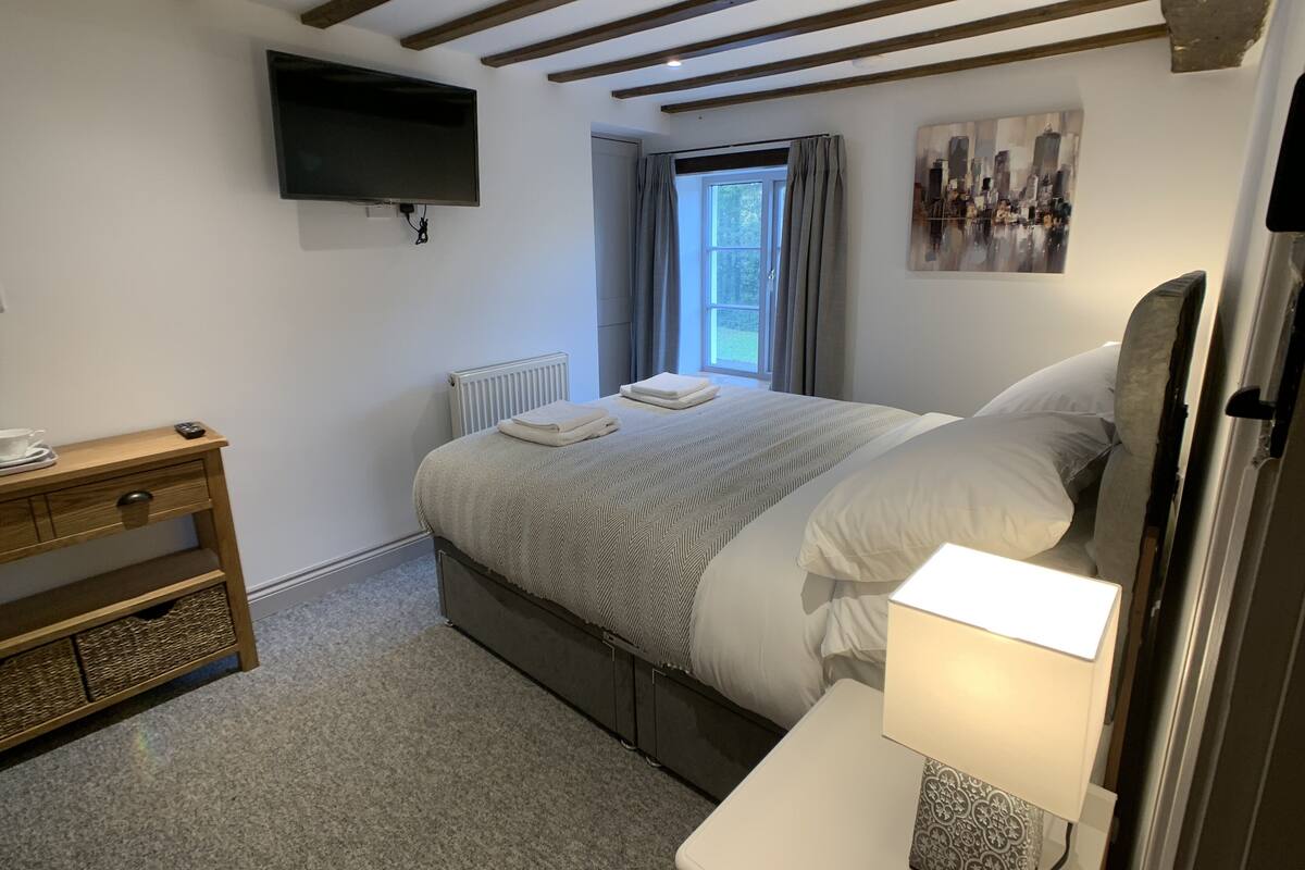 A bedroom with a bed, a bedside table, a lamp, a TV, a window with curtains, and a radiator.