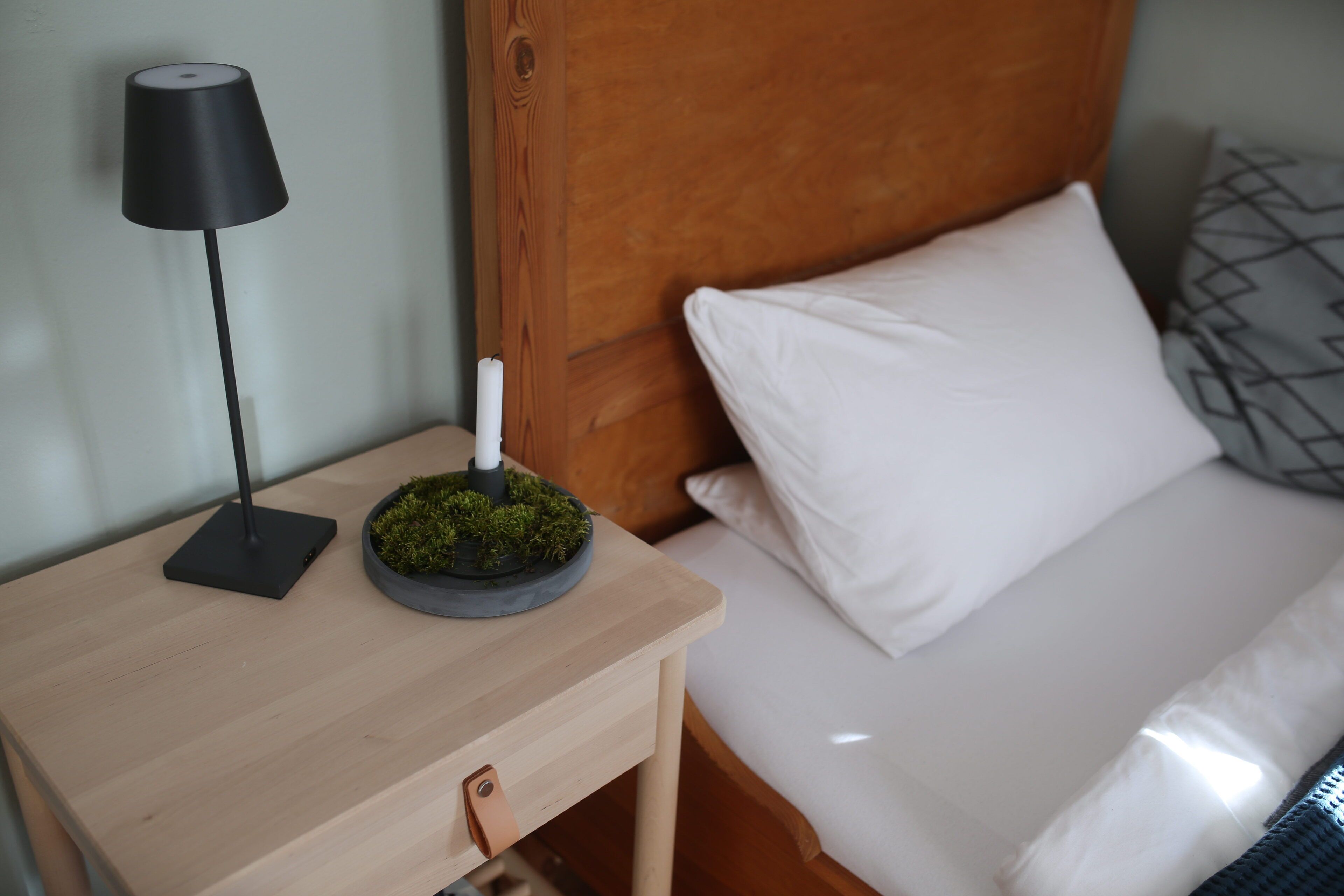 Una cama bien hecha con sábanas blancas, una mesita de noche de madera con una lámpara negra y una pequeña planta musgosa sobre la mesita.