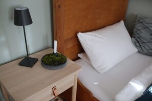 Een net opgemaakt bed met witte lakens, een houten nachtkastje met een zwarte lamp en een klein mosplantje op het nachtkastje.