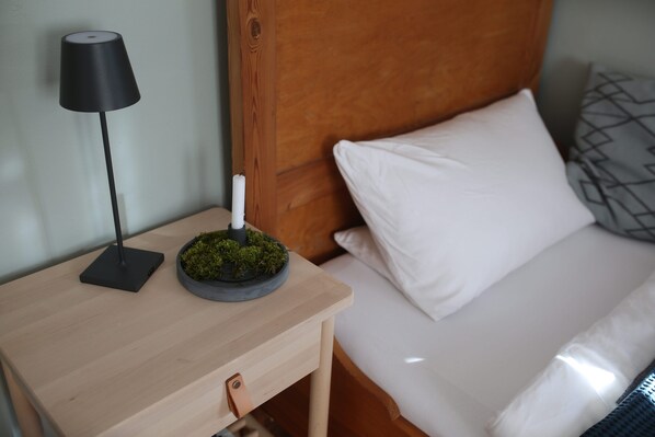 En pænt redt seng med hvide sengetæger, et trænatbord med en sort lampe og en lille mosplante på natbordet.