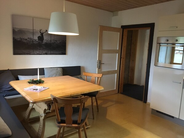 Een gezellige zithoek met een houten eettafel, twee stoelen, een bank en een groot schilderij. Een witte hanglamp verlicht de tafel.