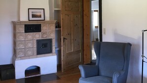 A room with a wooden ceiling, a blue armchair, a fireplace with a decorative cabinet, and a door leading to another room. - PrienZeit vacation apartment with fantastic panoramic views (Aschau)