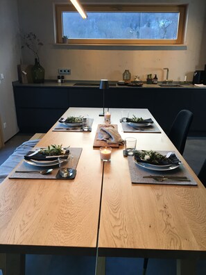 Une table à manger dressée avec des assiettes, des couverts, des verres et des bougies, avec une fenêtre au-dessus et une cuisine en arrière-plan.