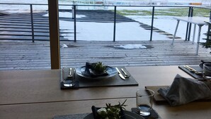 Een gedekte eettafel met borden, bestek en glazen, met uitzicht op een besneeerd landschap met bergen en een waterpartij.