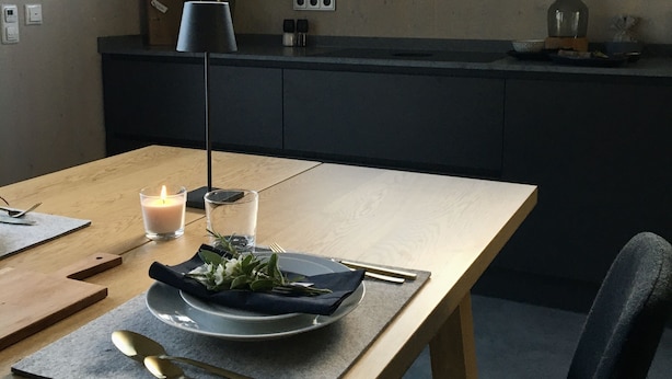 Een eetruimte met een houten tafel gedekt voor twee personen, een kaars, en uitzicht op bergen door een raam.