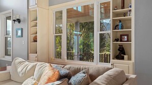 Uma sala de estar com um sofá bege, uma janela com vista para árvores e uma estante com diversos objetos.