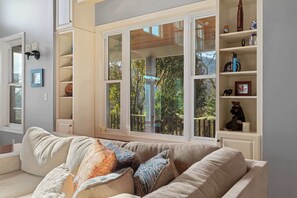Una sala de estar con un sofá color beige, una ventana que da a vista de árboles, y una estantería con diversos objetos.
