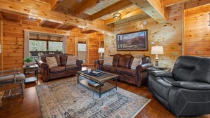 A cozy log cabin interior with wooden walls, a fireplace, leather sofas, and a large window offering a view of the outdoors. - W/C Accessible, Private Log Cabin, with Mountain Views, Private Hot Tub (Sevierville)