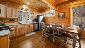 A wooden cabin-style kitchen with wooden cabinets, a stainless steel refrigerator, a dining table with chairs, and a window with a view of trees. - W/C Accessible, Private Log Cabin, with Mountain Views, Private Hot Tub (Sevierville)