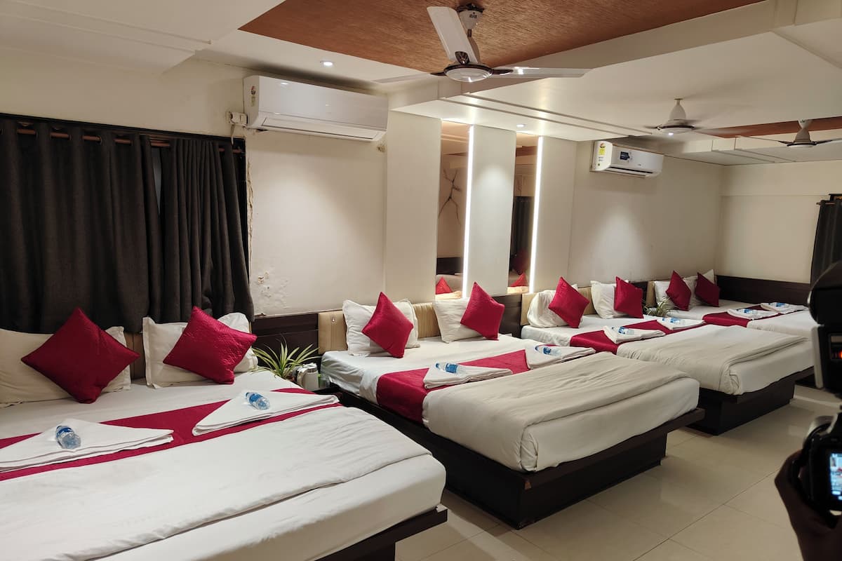 A hotel room with four beds arranged in a row, each with white bedding and red pillows. There are ceiling fans, air conditioning units, and a large mirror on the wall.