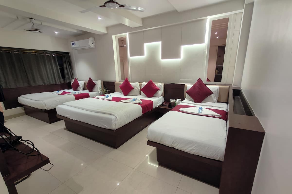 Habitación de hotel con cuatro camas individuales, cada una con ropa de cama blanca y almohadas rojas. Hay un ventilador de techo, un equipo de aire acondicionado y un espejo grande en la pared.