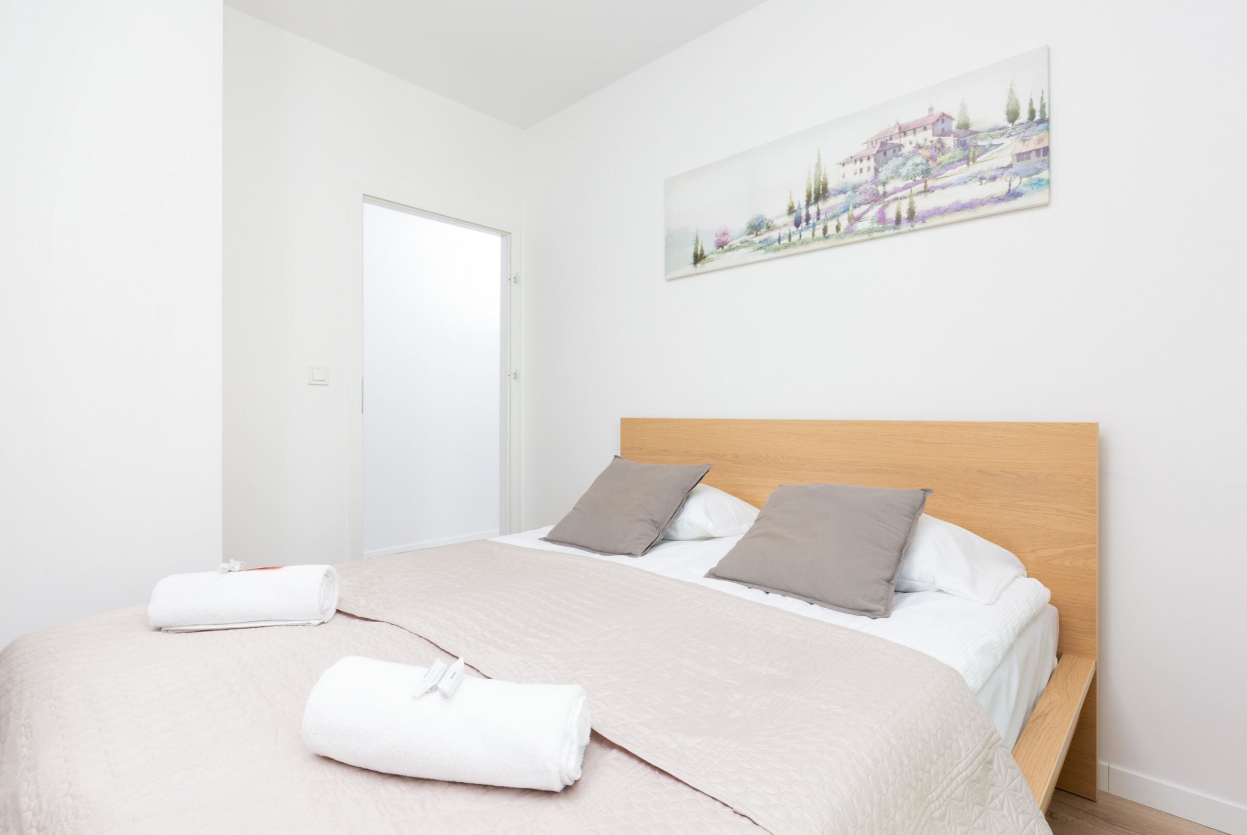 Un lit bien fait, avec une tête de lit en bois, deux oreillers et des serviettes roulées, posés sur un couvre-lit blanc, dans une pièce aux murs blancs et dotée d’une porte.