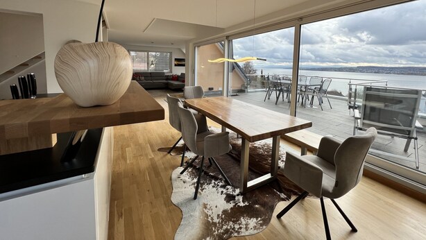 Ein moderner Essbereich mit Holztisch und -stühlen, einer großen Vase und Blick auf ein Gewässer durch große Fenster.