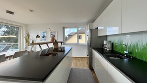 Eine moderne Küche mit weißen Schränken, einer schwarzen Arbeitsplatte und Blick auf ein gelbes Haus durch das Fenster.