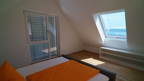 Ein Schlafzimmer mit schrägen Wänden, einem Bett mit orangefarbener Bettdecke, einem Fenster mit Jalousien und Blick ins Freie.