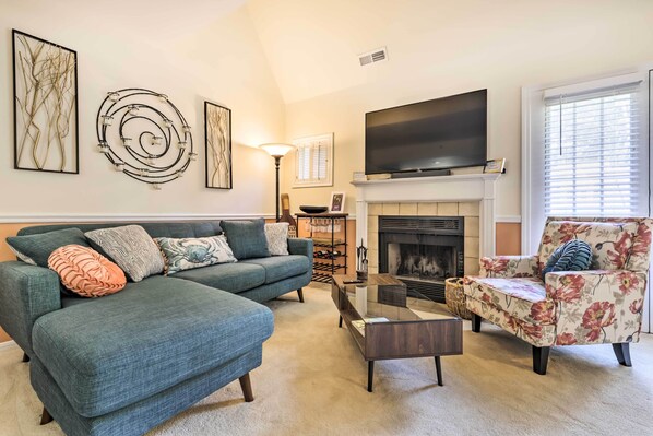 A living room with a sectional sofa, a floral armchair, a glass coffee table, and a fireplace. - Lovely Norwich Villa, On Golf Course w/ Amenities! (Norwich)