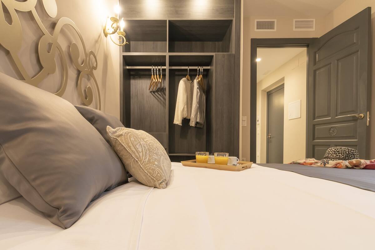 Una cama bien hecha con almohadas, una mesita de noche con un trayador de zumo de naranja y un armario con ropa colgada.