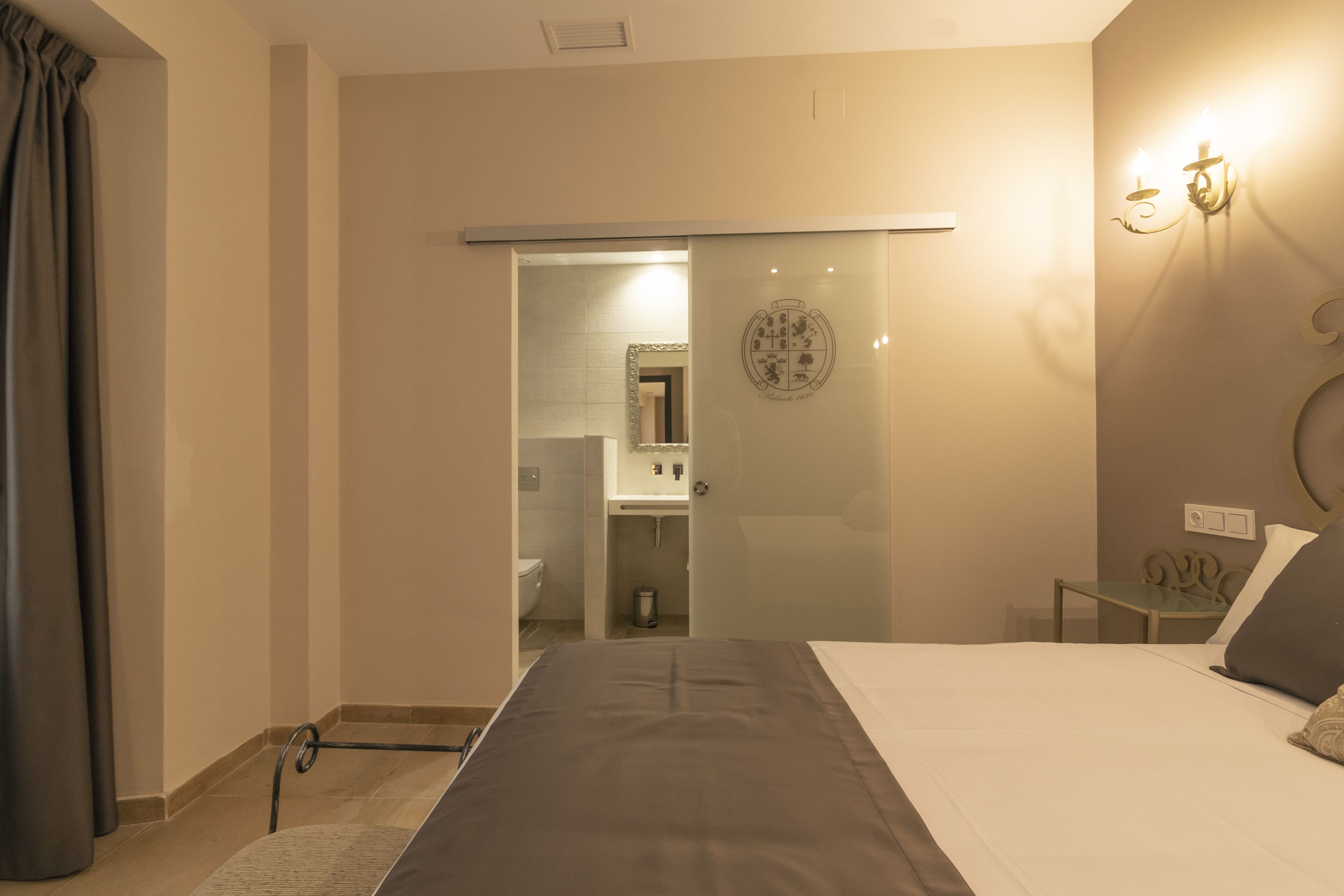 Una habitación de hotel con cama, mesita de noche, espejo y baño visible a través de una puerta de cristal.