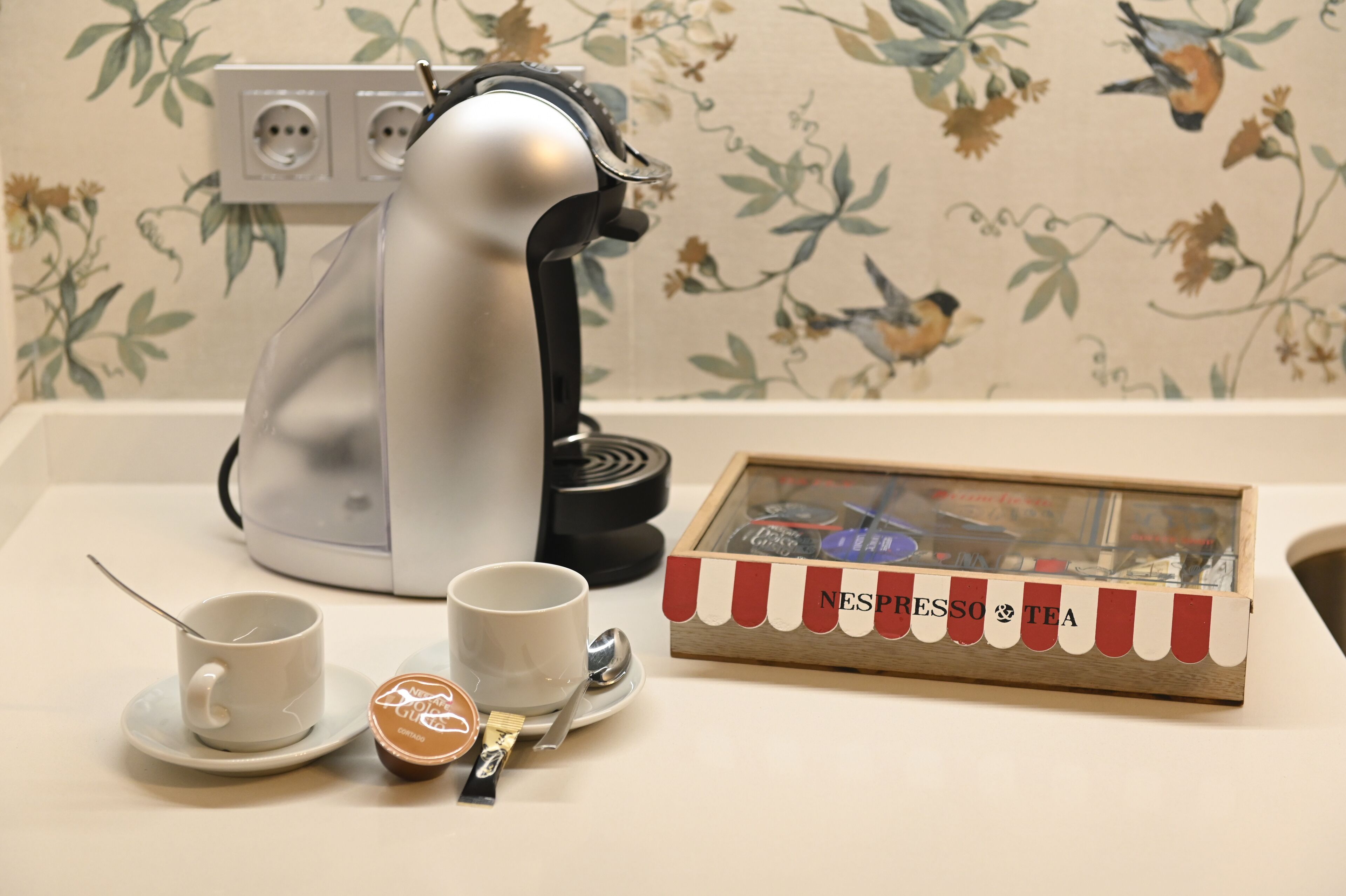 Una cafetera, dos tazas, una cuchara, un recipiente pequeño y una caja con la etiqueta 'NESPRESSO & TEA' sobre una mesa.
