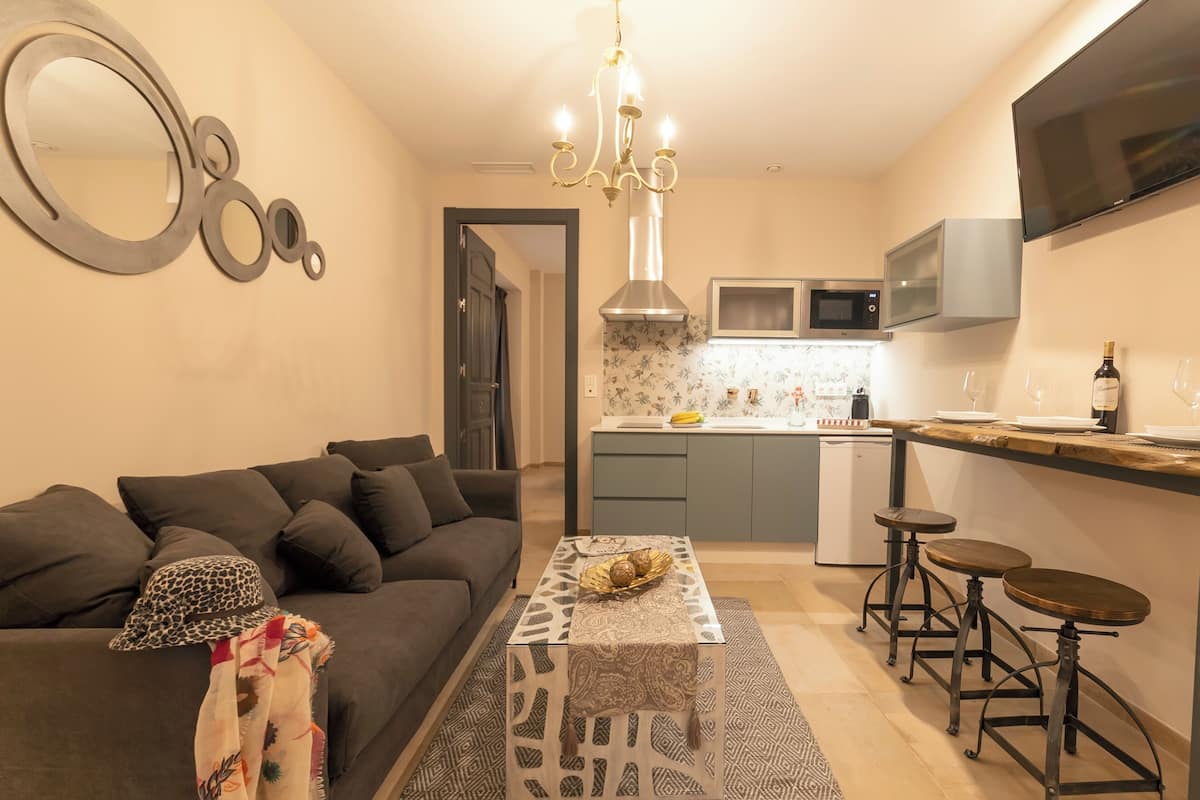 Un espacio habitable compacto con un sofá, una mesa de centro estampada, una araña, una cocina con microondas y un televisor montado en la pared.