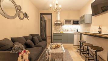 A compact living space with a sofa, a patterned coffee table, a chandelier, a kitchen with a microwave, and a TV mounted on the wall.