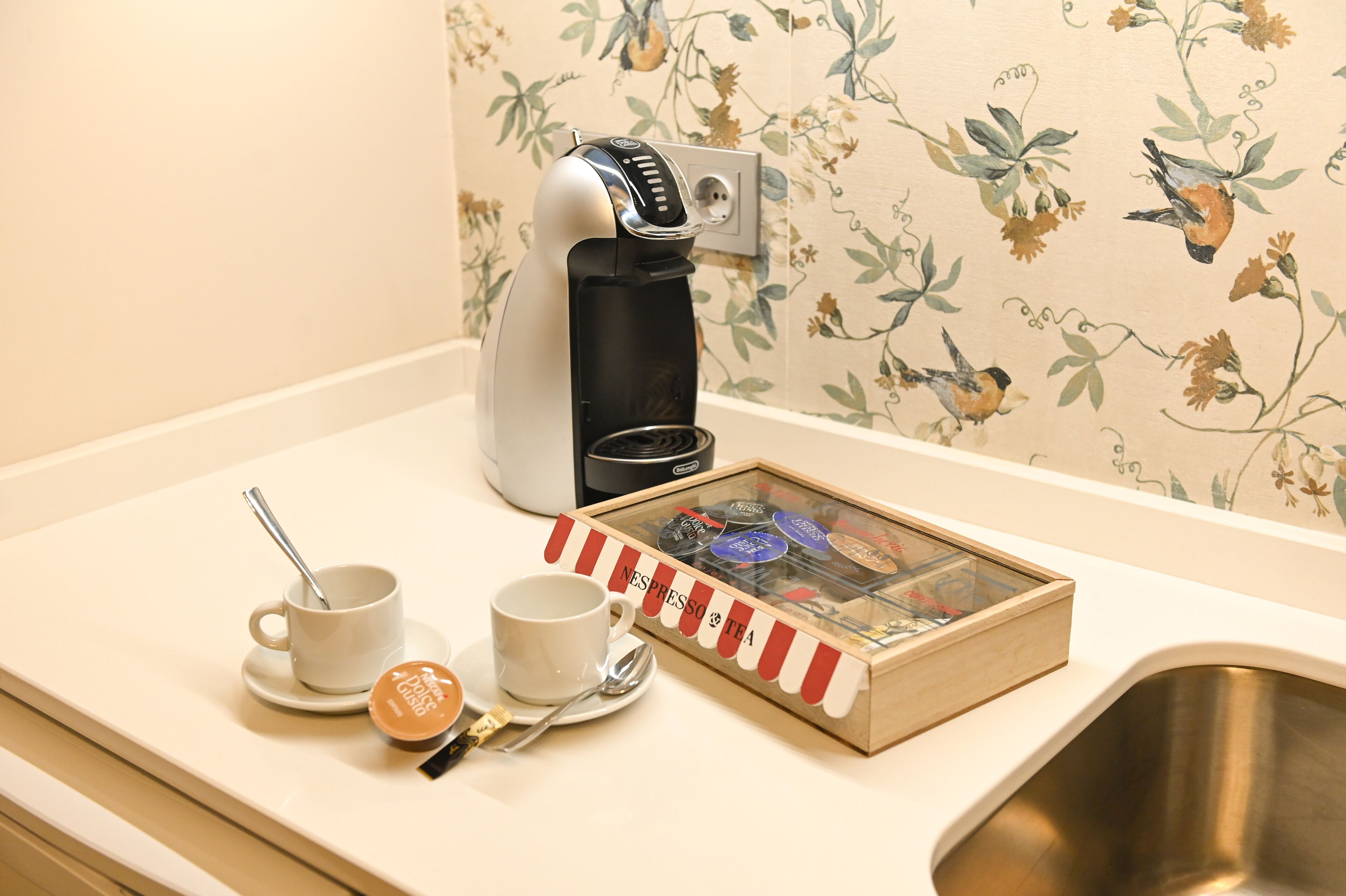 Une machine à café, deux tasses, une cuillère et une boîte de sachets de thé Nespresso sont posés sur un comptoir.